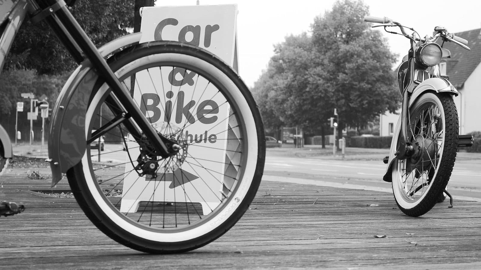 Motorrad-Vorderrad vor dem Car-&-Bike-Fahrschulschild – Schwarz-Weiß-Foto
